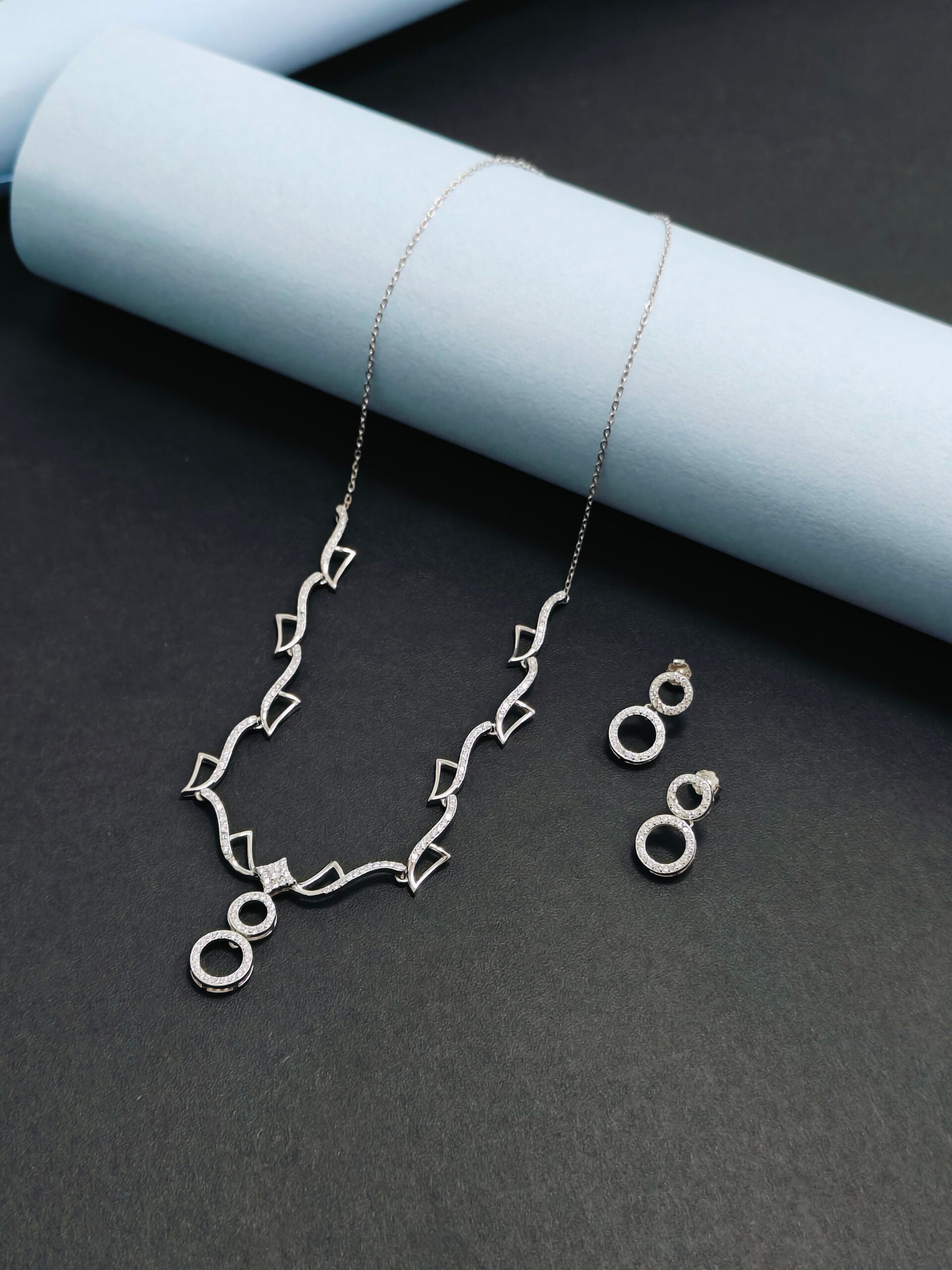 Silver necklace and earrings on a dark surface with light blue cylindrical objects.